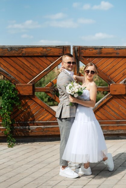 Noiva jovem em um vestido curto branco e noivo em um terno cinza perto do portão de madeira
