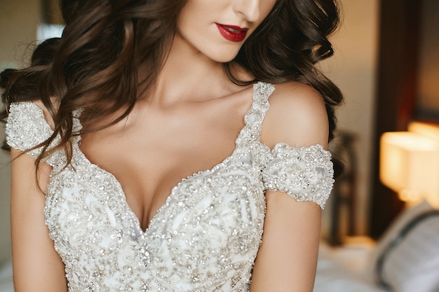 Noiva linda com maquiagem de casamento e cabelos longos ondulados em vestido de noiva. modelo de vestido de noiva elegante posando no interior. jovem mulher em um vestido de luxo decorado com cristais. moda casamento