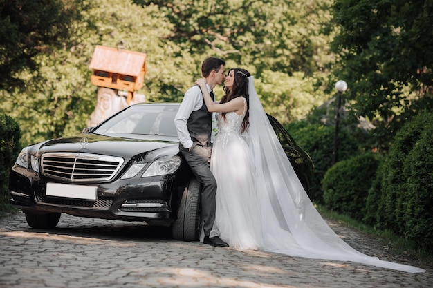 Noiva linda com maquiagem e penteado à moda em vestido de noiva de luxo com noivo bonito perto de carro de casamento preto