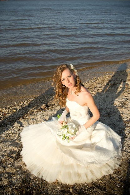 Noiva linda em um vestido branco na costa do rio no verão. Maquiagem e penteado profissionais