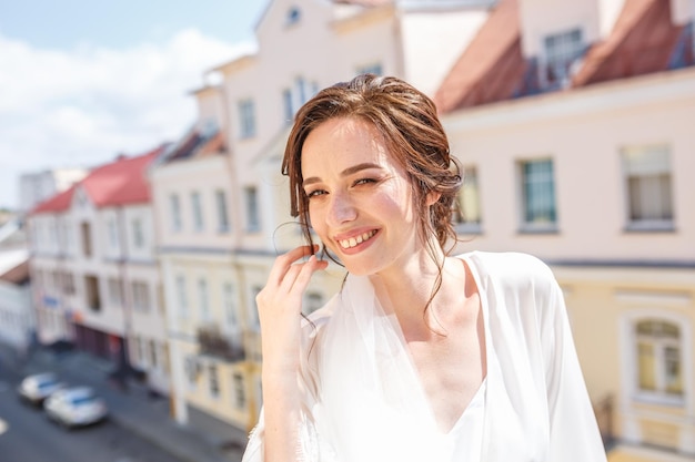 Noiva linda jovem em um peignoir em pé na varanda com vista para a cidade velha e esperando o noivo