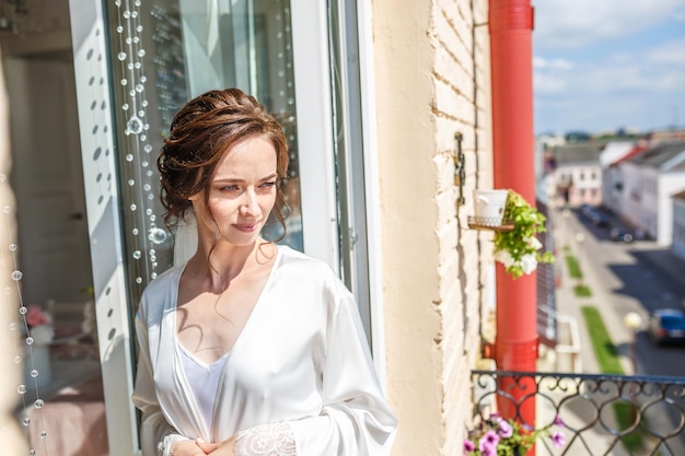 Noiva linda jovem em um peignoir em pé na varanda e esperando o noivo