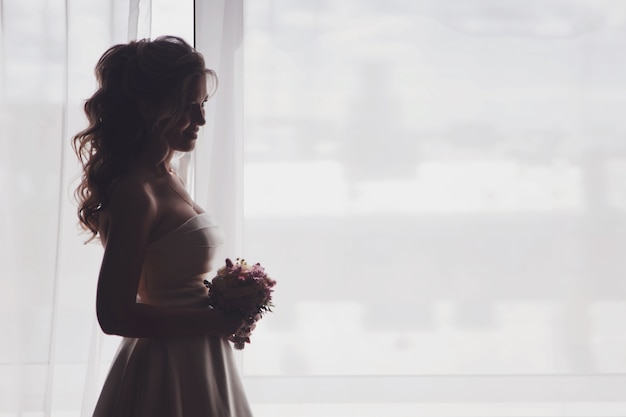 Noiva linda jovem num vestido com buquê de flores na janela em quarto de hotel. Manhã da noiva no dia do casamento. Mulher feliz está esperando para conhecer o noivo. Conceito de feliz e luxo casado. Copie o espaço