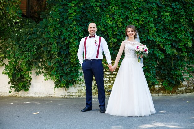 Noiva lindo casal em um vestido branco com um buquê, enquanto o noivo com suspensórios e gravata borboleta de mãos dadas. no contexto de uma parede com folhas verdes. casal feliz. conceito de casamento.
