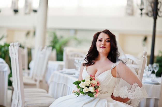 Noiva morena de seios grandes com buquê sentado na cadeira no salão de casamento
