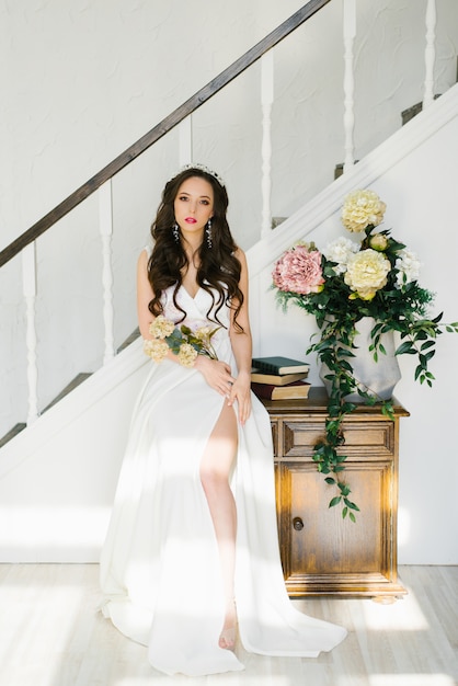 Noiva morena linda em um vestido de noiva branco com uma fenda na perna em um interior clássico. Ela segura nas mãos as flores