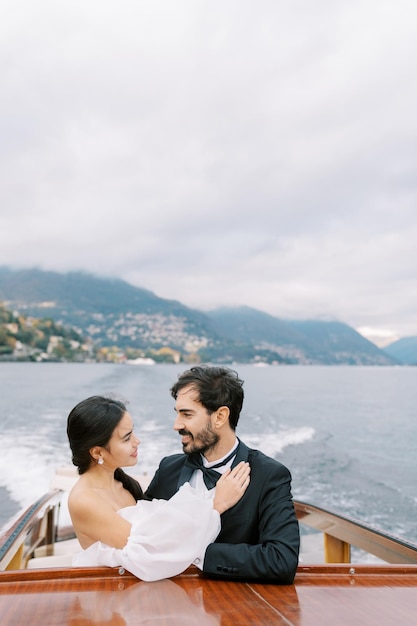 Noiva sorridente toca o peito do noivo enquanto está de pé em um iate navegando no lago como Itália