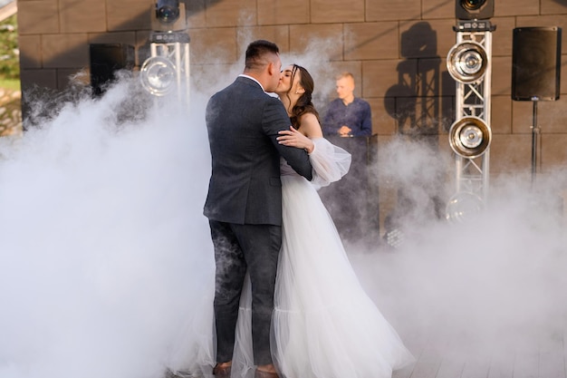 Noivas amorosas se beijando em cena com fumaça tendo a primeira dança de casamento Belo momento para a garota do casal em lindo vestido de casamento inchado com noivo de terno comemorando