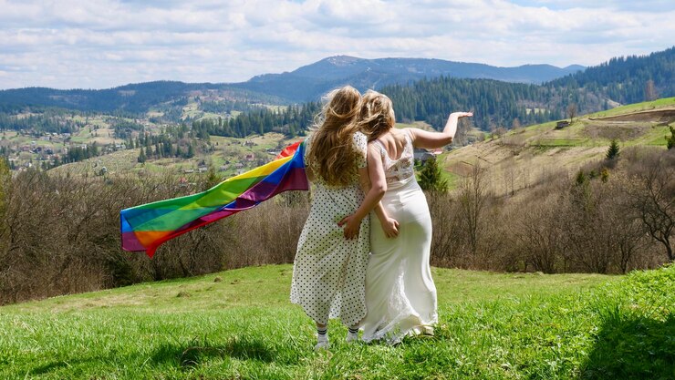 Foto noivas lésbicas mulheres loiras casadas em vestidos de noiva brancos com bandeira lgbt ao ar livre nas montanhas garotas homossexuais de costas abraçadas pela bunda sem rosto