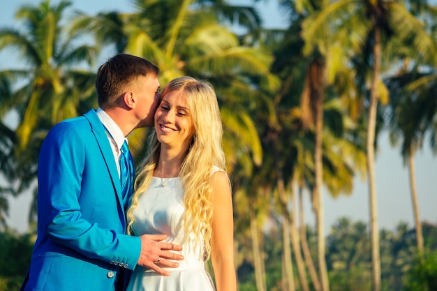 Noivo bonito em um terno de luxo e uma linda noiva em um lindo vestido de noiva, sorrindo e posando em um fundo de palmeiras. conceito de uma cerimônia de casamento chique e rica na praia