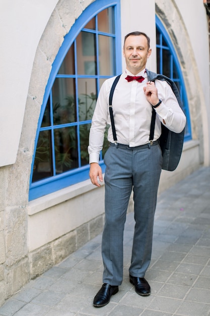 Noivo de homem caucasiano maduro bonito em uma camisa branca com uma gravata borboleta vermelha e suspensórios segura uma jaqueta, posando em frente a um edifício vintage com uma grande janela azul ao ar livre na cidade