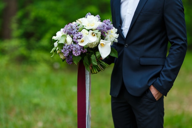 Noivo de terno azul escuro segurando um grande buquê de casamento lilás e fitas com fundo de jardim