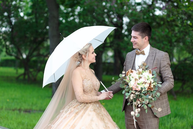 Noivo e noiva felizes com um guarda-chuva branco na chuva, no verão no parque. Casamento ao ar livre.