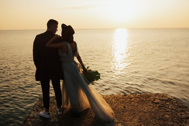 noivo e noiva olham para o lindo pôr do sol laranja perto do mar