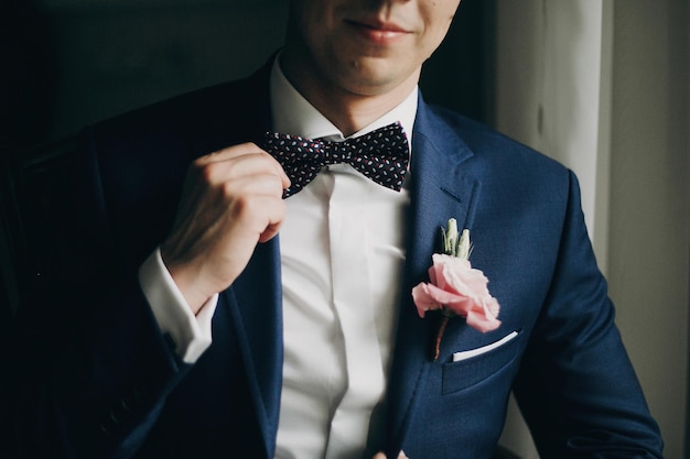 Noivo elegante de terno azul com flor na lapela com rosa segurando gravata borboleta e posando perto da janela no quarto de hotel Preparação matinal antes da cerimônia de casamento