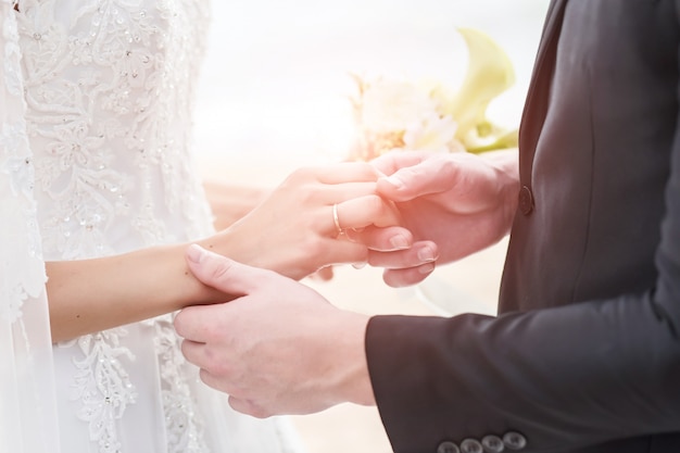 Noivos que guardam as mãos com alianças de casamento no fundo do mar e do sol.