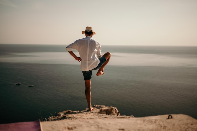 Un nómada digital, un hombre con sombrero, un empresario hace yoga en las rocas junto al mar al atardecer hace un