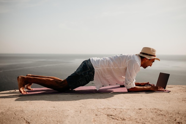 Un nómada digital, un hombre con sombrero, un empresario con un portátil hace yoga en las rocas junto al mar en