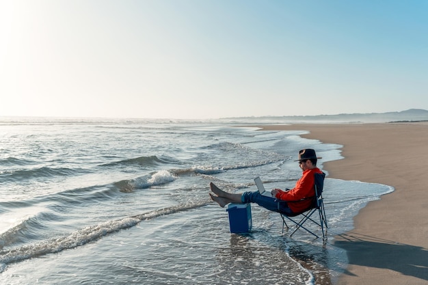 Nómada digital sentado con una laptop en la orilla de la playa solo trabajando relajado