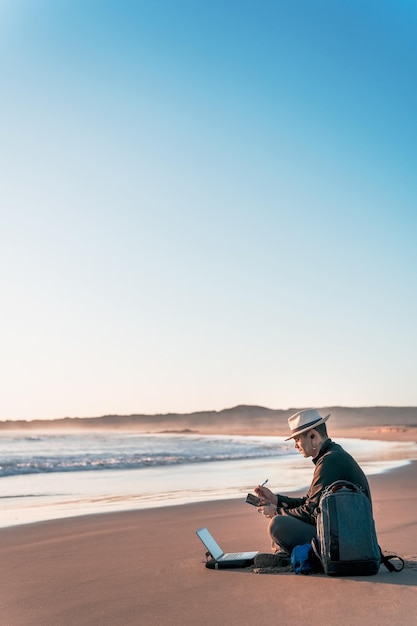 Nómada digital sentado en la playa con su computadora portátil y tomando notas en la orilla de la playa al atardecer