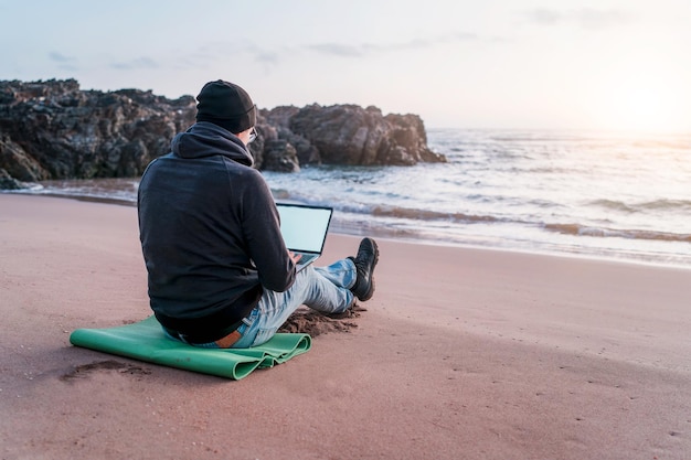 Nómada digital trabajando en su computadora portátil al aire libre desde la playa al atardecer vista trasera