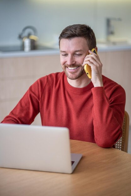 Nômade digital satisfeito homem freelancer sorrindo conversa celular chamada remota trabalhando com laptop em casa