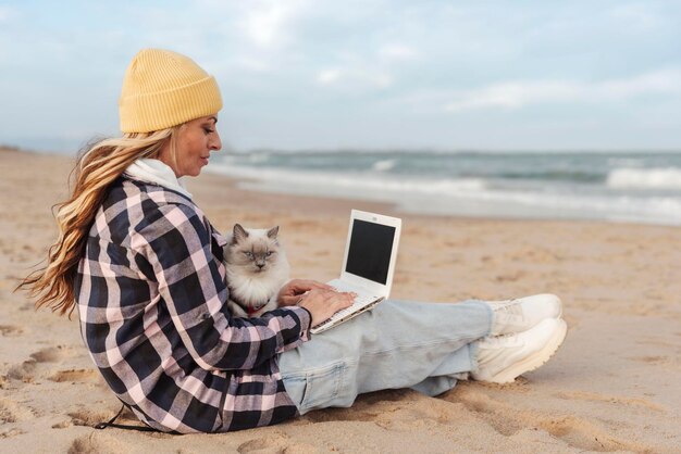 Nomadenfrau mit Laptop-Computer an einem Strand, der mit ihrer Katze sitzt Freiberufliches Arbeitskonzept