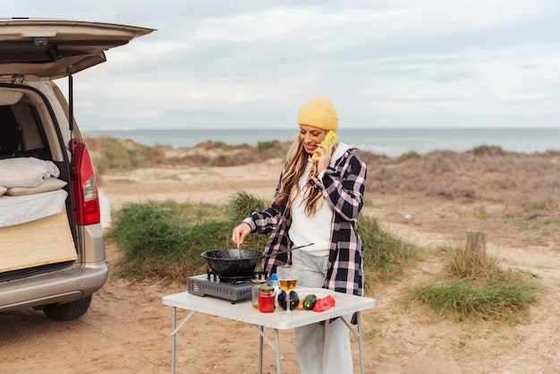 Nomadenreisende Frau spricht per Handy, während sie außerhalb des Wohnmobils neben dem Strand kocht