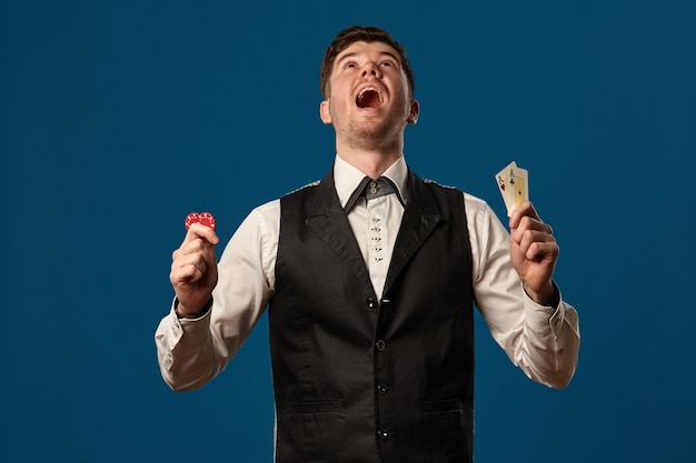 Noob im Poker in schwarzer Weste und weißem Hemd mit zwei roten Chips und Assen posiert vor blauem Hinterg...