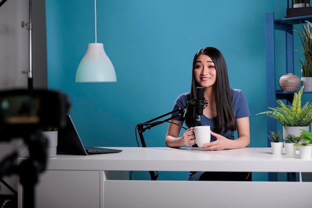 Nos bastidores do videoblogueiro asiático sentado na mesa em frente ao podcast de gravação da câmera para o canal de vlogging em estúdio com fundo azul. Influenciador de mídia social usando equipamento de produção