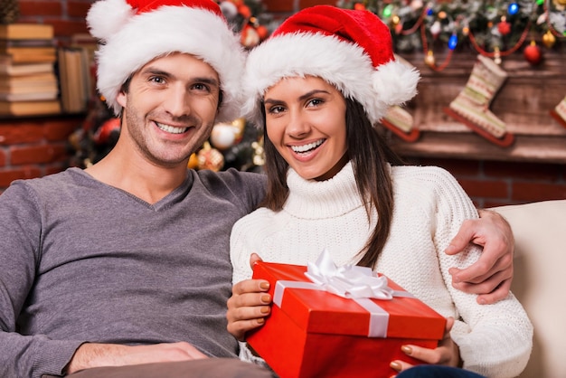 ¡Nos encanta la Navidad! Hermosa joven pareja amorosa pegado entre sí y sonriendo mientras mujer sosteniendo caja de regalo con árbol de Navidad en el fondo