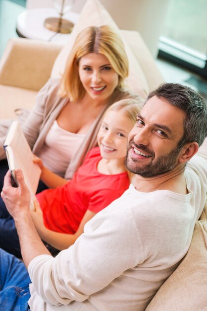 Nos encanta pasar tiempo juntos. Vista superior de la familia feliz de tres uniéndose entre sí y sonriendo mientras está sentado en el sofá y lee un libro juntos