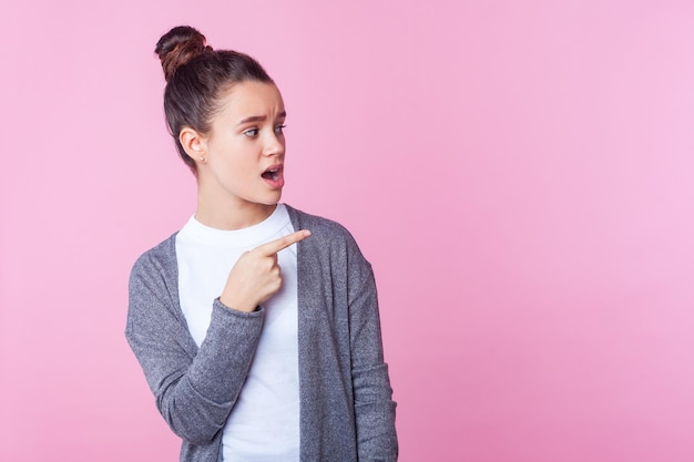 Nossa, anuncie aqui. Retrato de adolescente maravilhado com penteado de coque em roupas casuais, olhando com a boca aberta de surpresa, apontando para o lado. tiro de estúdio interior isolado no fundo rosa