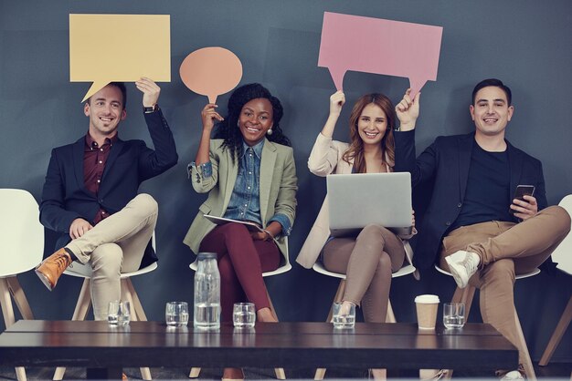 Nossas opiniões nos levarão ao sucesso Foto de um grupo de empresários segurando balões de fala e usando diferentes dispositivos sem fio enquanto esperam na fila para uma entrevista