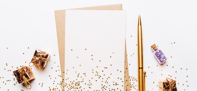 Nota en blanco con sobres, regalos, bolígrafo y estrellas de brillo dorado sobre fondo blanco.