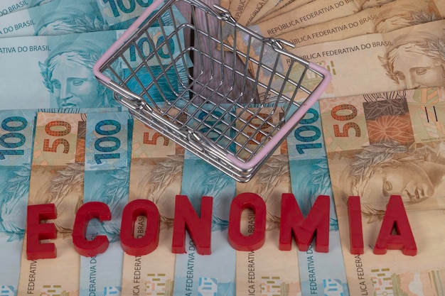 Notas brasileiras Notas de 50 e 100 reais ao fundo com a palavra economia em economia portuguesa em inglês em vermelho com cesta de mini supermercado Foco seletivo