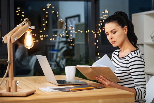 Notas importantes. Mujer agradable seria inteligente sosteniendo un cuaderno y leyendo sus notas mientras trabaja hasta tarde en la oficina