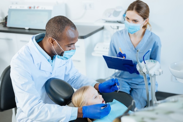 Foto notas vitais. jovem e charmosa enfermeira fazendo anotações das observações do dentista enquanto ele faz um exame completo da cavidade oral de uma garotinha