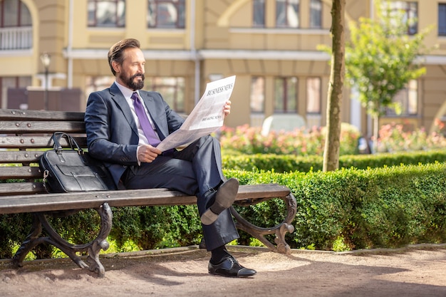 Notícias diárias. Comprimento total de um empresário sorridente e agradável lendo um jornal enquanto está sentado no banco
