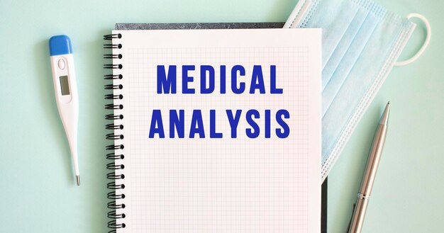 Notizblock medizinische Maske Thermometer und Stift auf blauem Hintergrund MEDIZINISCHER ANALYSE-Text in einem Notizbuch