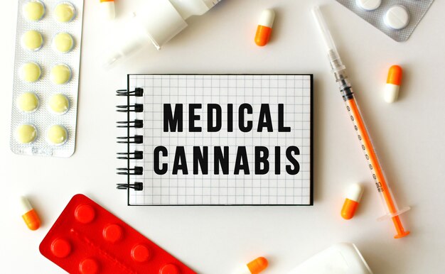 Notizblock mit Text MEDICAL CANNABIS auf weißem Hintergrund. In der Nähe befinden sich verschiedene Medikamente. Medizinisches Konzept.