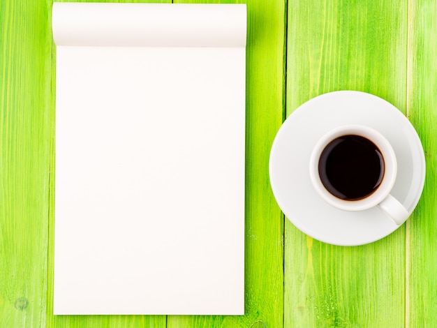Notizblock offen mit weißer Leerseite für das Schreiben von Idee oder von Aufgabenliste, Tasse Kaffee auf grüner hölzerner Tabelle