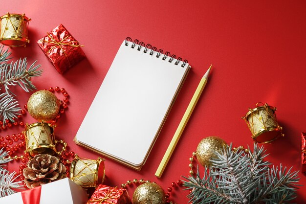 Notizblock und Bleistift zum Schreiben von Wünschen und Geschenken für das neue Jahr und Weihnachten um die Christbaumschmuck auf einem roten Tisch. Von oben betrachten.