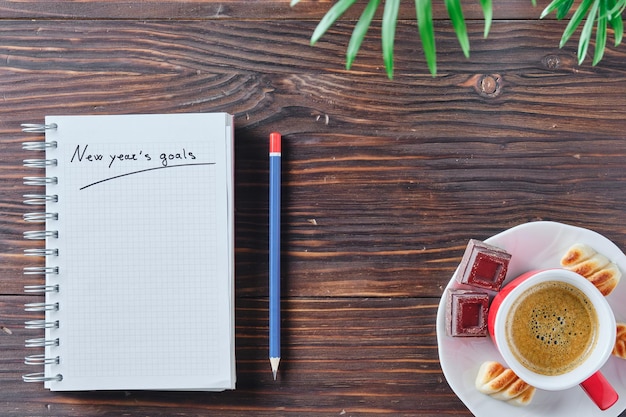 Notizbuch mit den Worten Neujahrsziele auf einem rustikalen braunen Holzhintergrund mit einem blauen und roten Bleistift daneben, einigen grünen Blättern oben und einer Tasse Kaffee in der Ecke