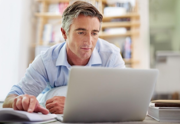 Notizen vergleichen Aufnahme eines gutaussehenden reifen Mannes, der mit einem Laptop auf dem Boden seines Wohnzimmers liegt