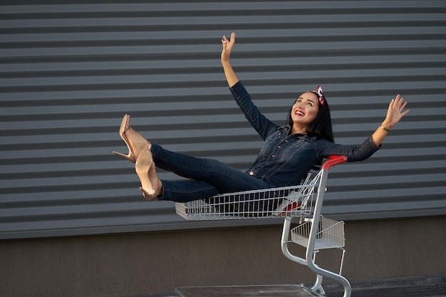 Nova coleção da moda. Retrato feliz feliz elegante menina morena com cabelos longos, vestindo macacões jeans da moda, montando um carrinho de compras. Filmado na parede urbana cinza