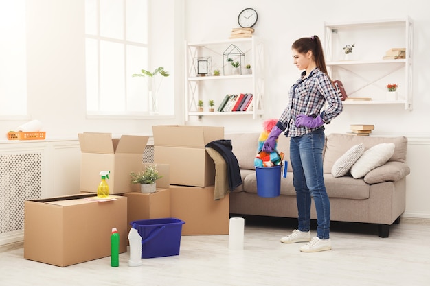 Nova limpeza doméstica. Jovem mulher melhorando depois de se mudar para um novo apartamento. Menina com vários detergentes, trapos e esfregões na sala cheia de caixas de papelão. Conceito de limpeza doméstica e de primavera