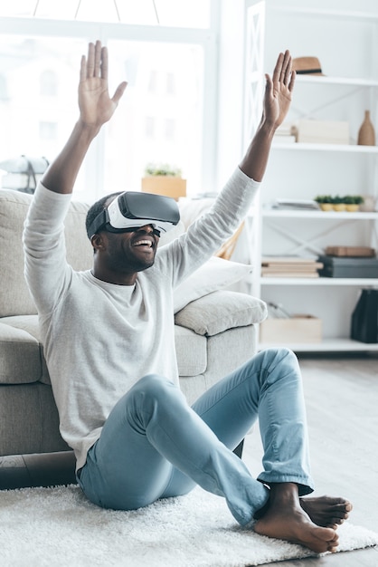 Nova palavra de realidade virtual! Jovem africano bonito com fone de ouvido VR gesticulando e sorrindo enquanto está sentado no tapete em casa