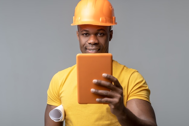 Novas tecnologias. jovem afro-americano sorridente atraente com capacete laranja e tablet na mão sobre fundo claro.