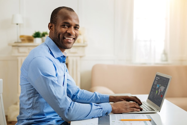 En la novena nube. Hombre afroamericano inspirado guapo sonriendo y trabajando en la computadora portátil mientras está sentado en la mesa